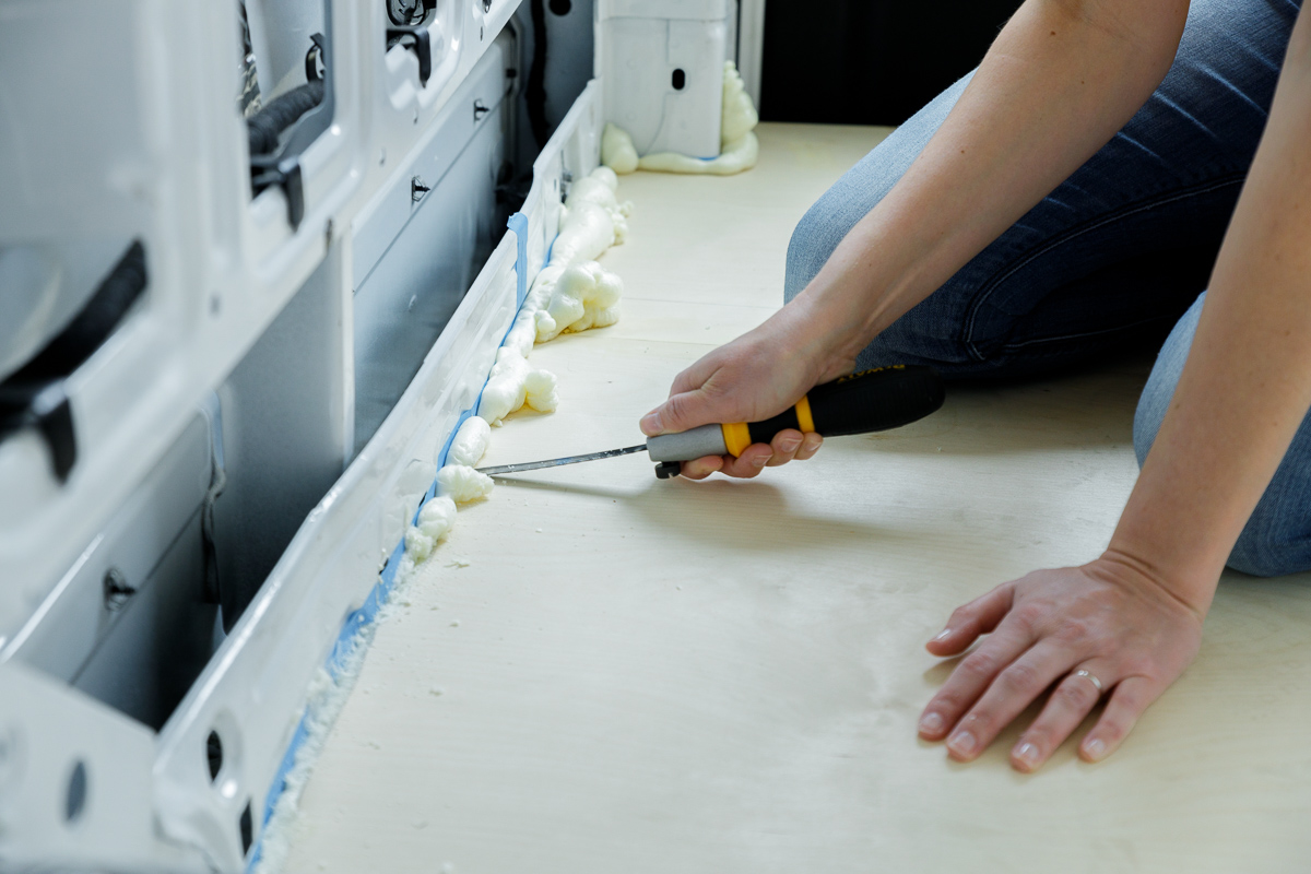 Close up on a woman's hands as she trims excess expanding foam from the perimeter of a plywood subfloor installed in the rear of a campervan