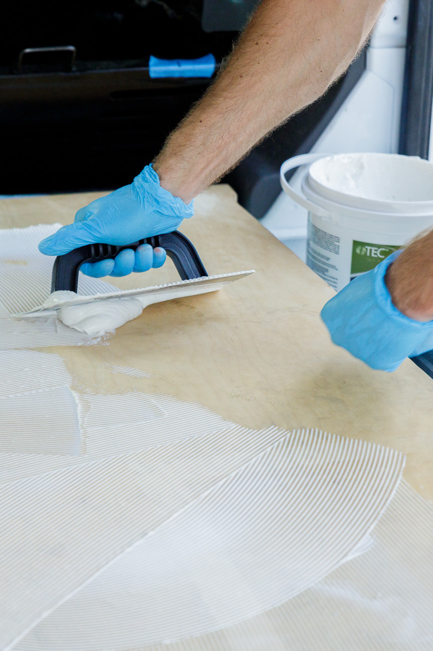 Close up on a man's gloved hands as he spreads sheet vinyl adhesive from a bucket onto the plywood subfloor of a campervan conversion 