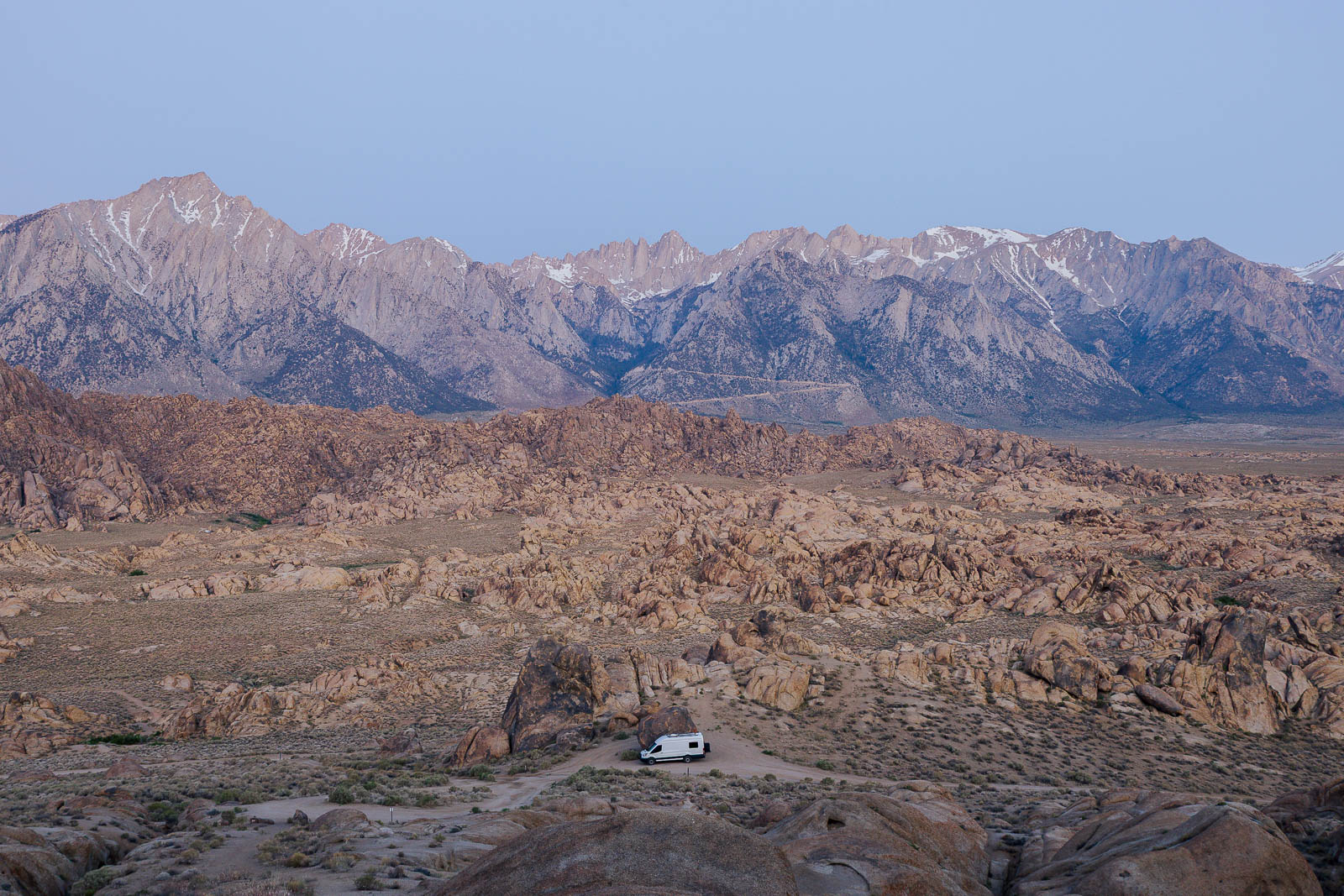 A white campervan parked among large boulders at Alabama Hills with the Sierra Nevada Mountains catching cool morning light at sunrise