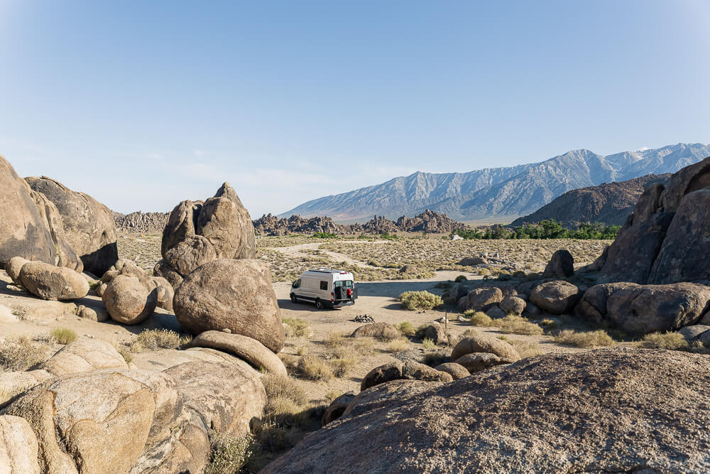 A campervan parked in a flat and secluded campsite surrounded by huge boulders with views of the Sierra Nevada Mountains in the distance