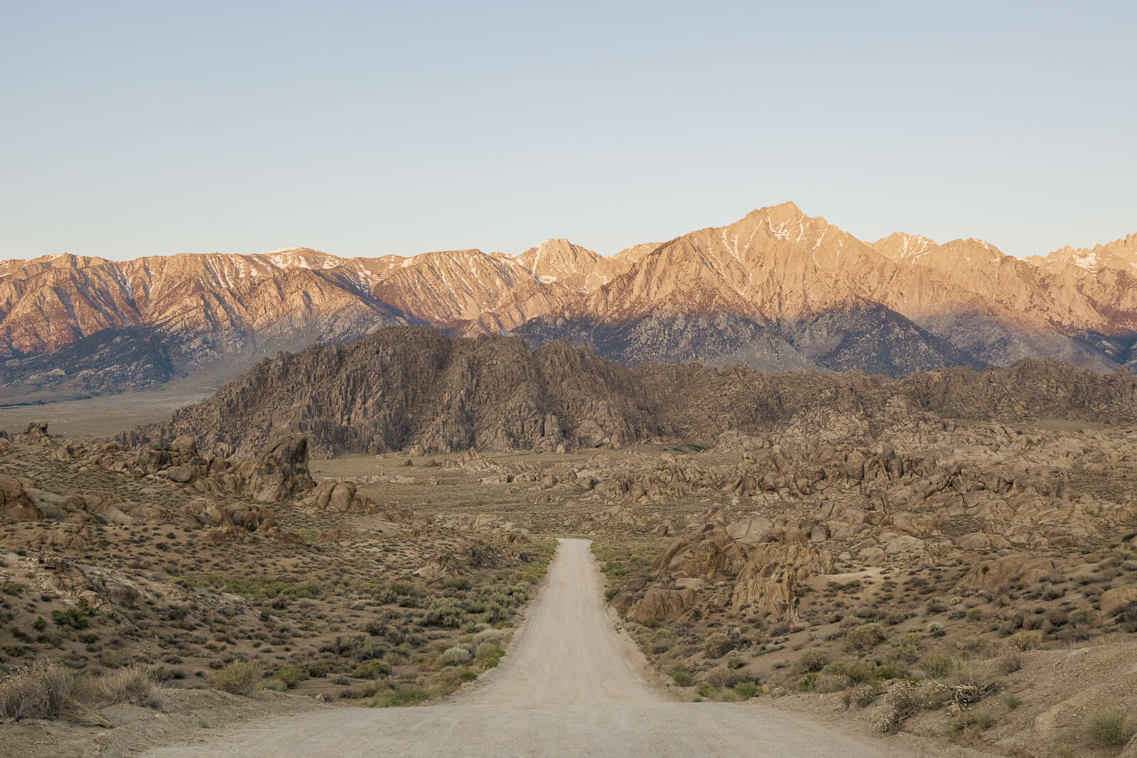 A dirt road leading toward a rocky landscape with the Sierra Nevada Mountains catching the warm light of sunrise in the distance