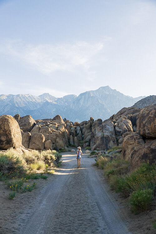 A woman standing in the middle of a narrow dirt road leading toward large boulders and distant mountains