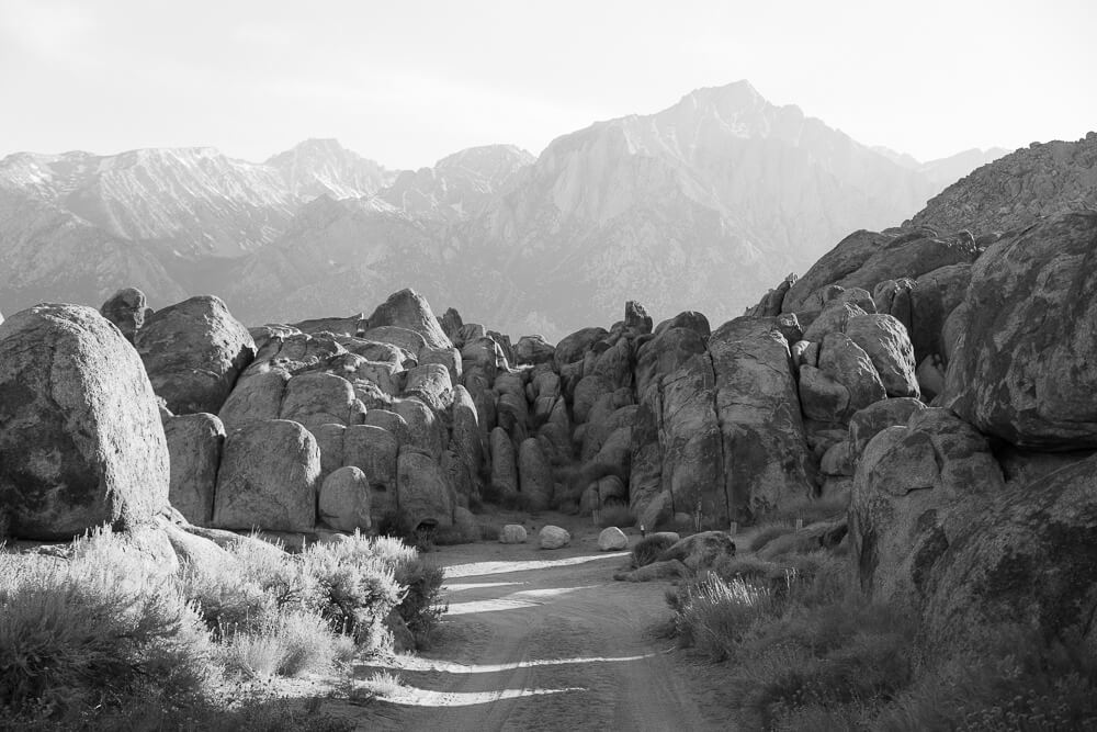 A black and white photo looking down a dirt road at large boulders with mountain peaks in the distance