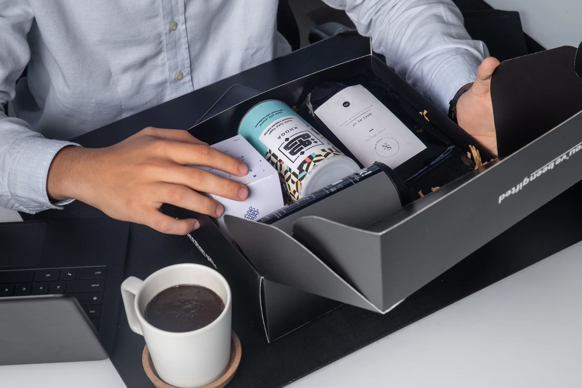 Person opening a black gift box containing a can, a pouch, and other packaged items on a desk with a laptop and a cup of coffee.