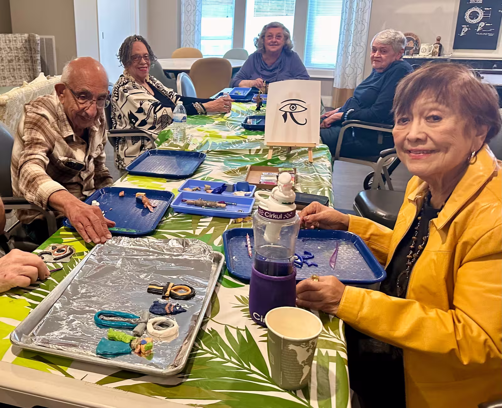 Seniors sitting around a table with blue trays and crafting materials, engaged in an art activity in a well-lit room with large windows. A small easel on the table displays an illustration of the Eye of Horus symbol.