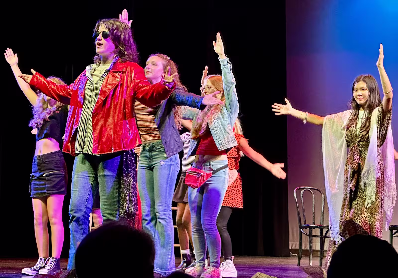 A group of young actors on stage performing a musical or play with one central actor wearing a red leather jacket and sunglasses, surrounded by others in casual costumes, all with their arms raised.
