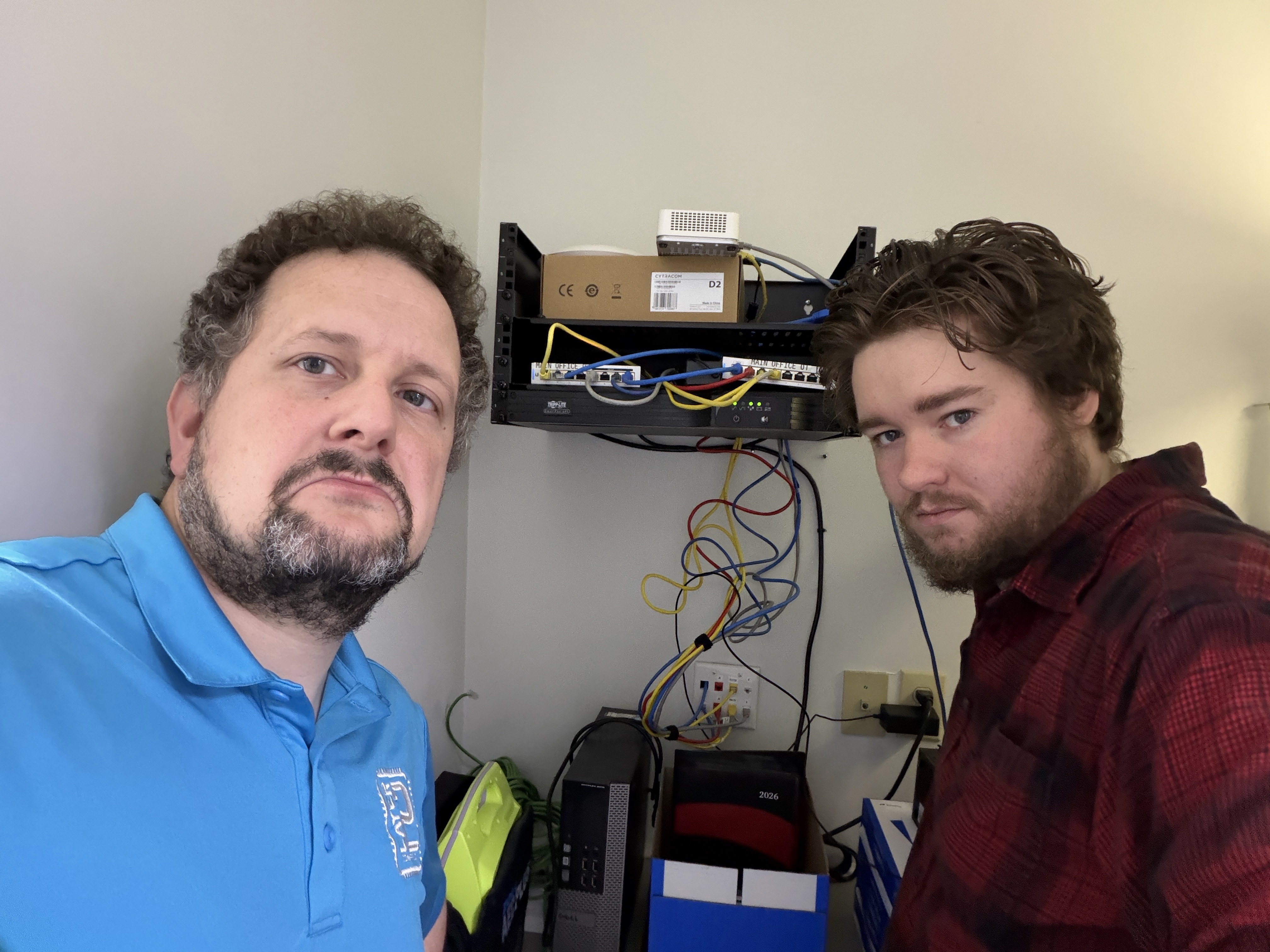 Layer 2 Computers technicians standing beside the original network rack and cluttered cabling before a small business network upgrade.