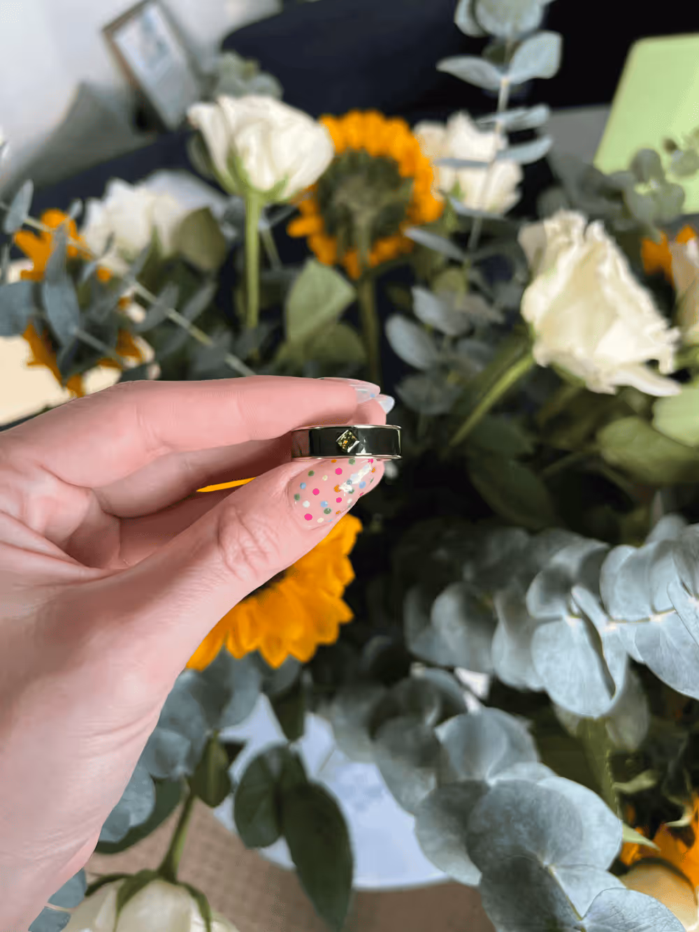 Hand with colorful polka dot nail art holding a silver ring with a small square yellow-green gemstone in front of a bouquet of sunflowers and white flowers.