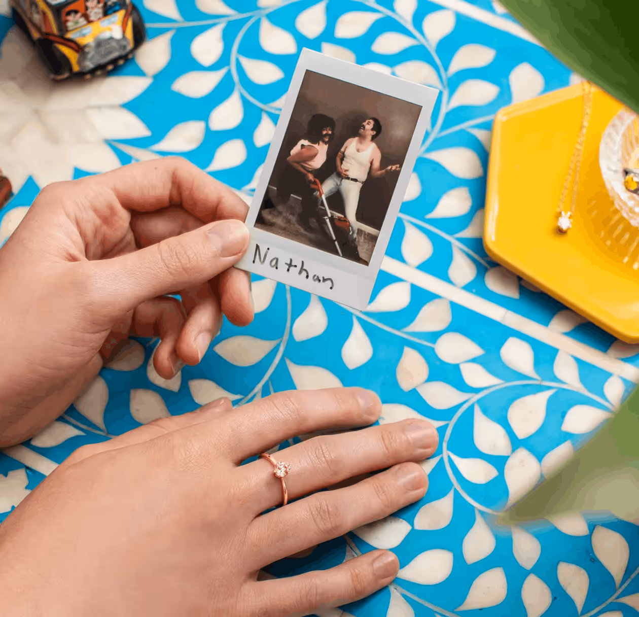 Person holding a Polaroid photo of two men posing with a vacuum cleaner, captioned 'Nathan', resting on a blue and white patterned table with a hand wearing a ring nearby.