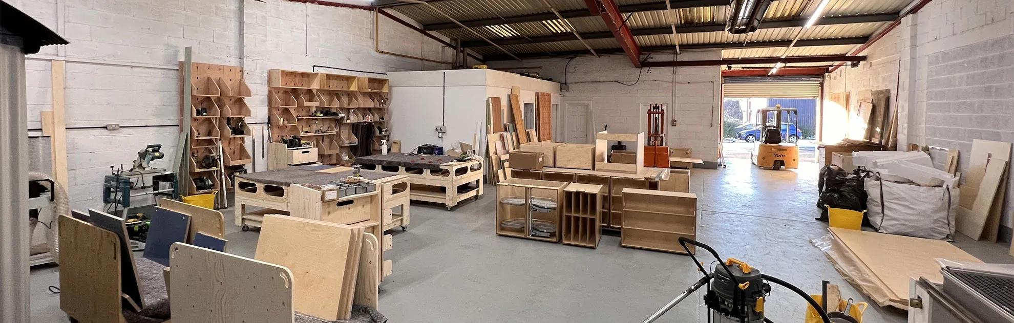Spacious woodworking shop with wooden shelves, workstations, plywood sheets, and a forklift near an open garage door.