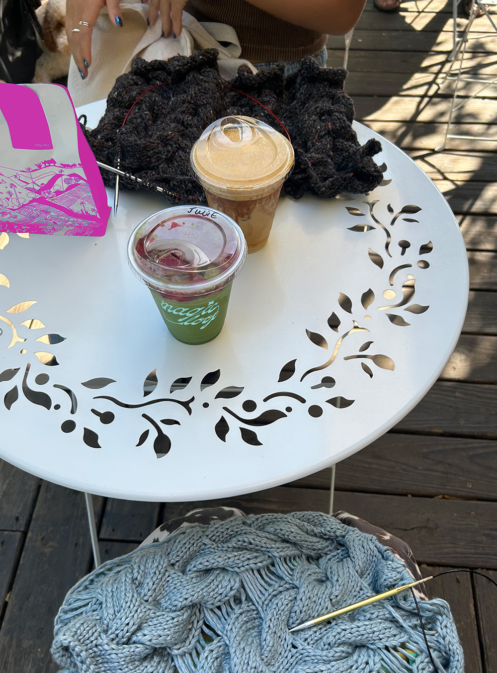 Magic Loop-branded coffee shop drinks on a white outdoor table with knitting projects.