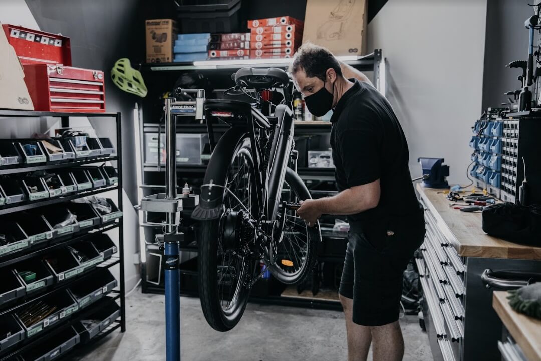 Man wearing a black face mask repairing a bicycle mounted on a repair stand in a workshop.