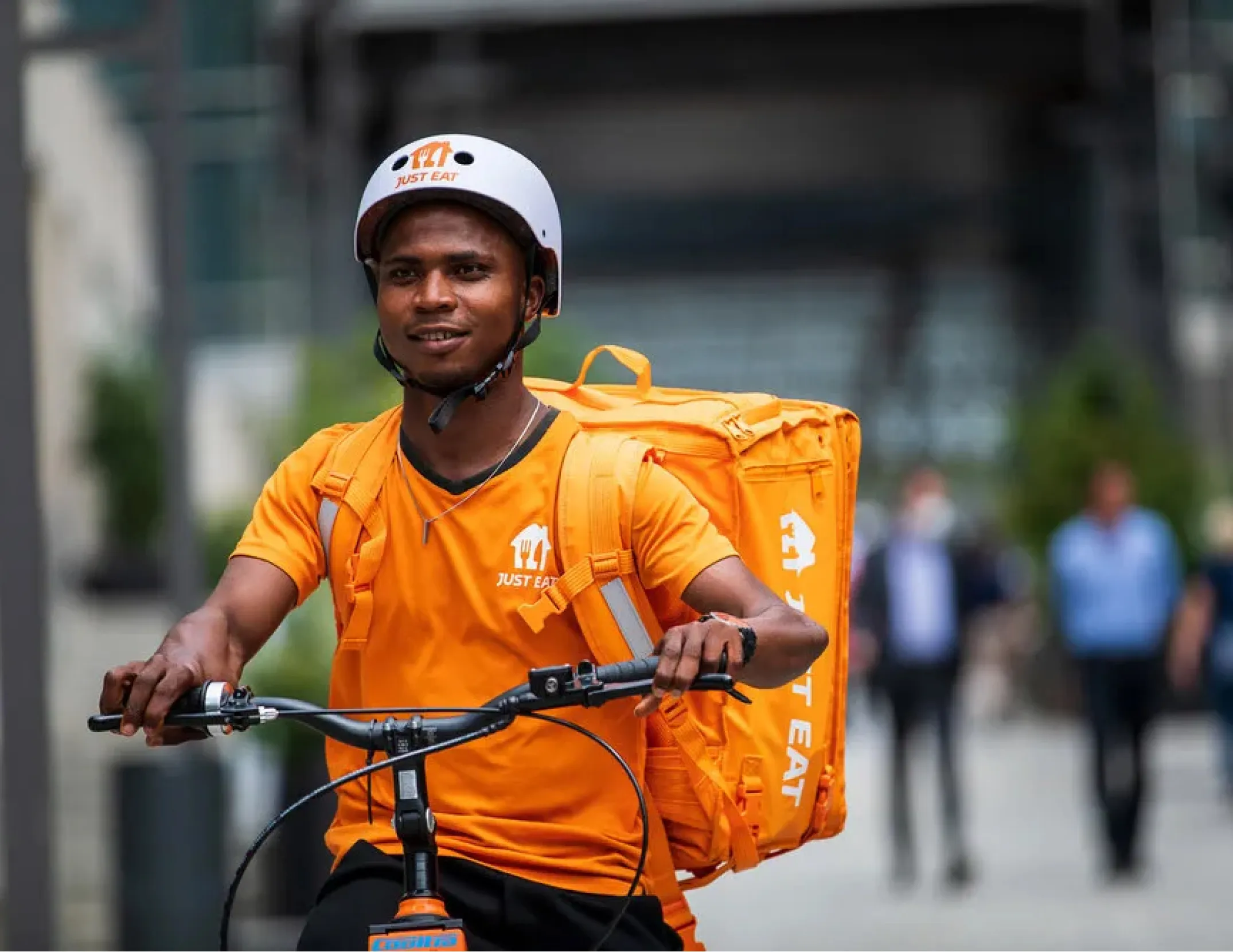 Woman in black jacket sitting on a white Zoomo electric bike with a large black backpack, in front of a brick and stone building.