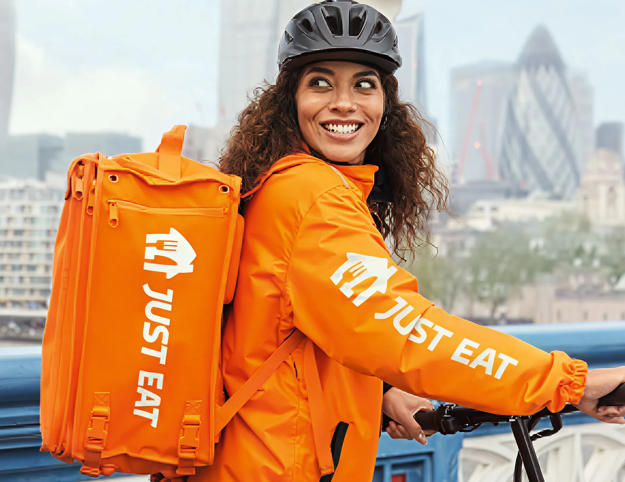 Man in black jacket and pants standing next to a black Zoomo electric bike against a brick and stone wall.