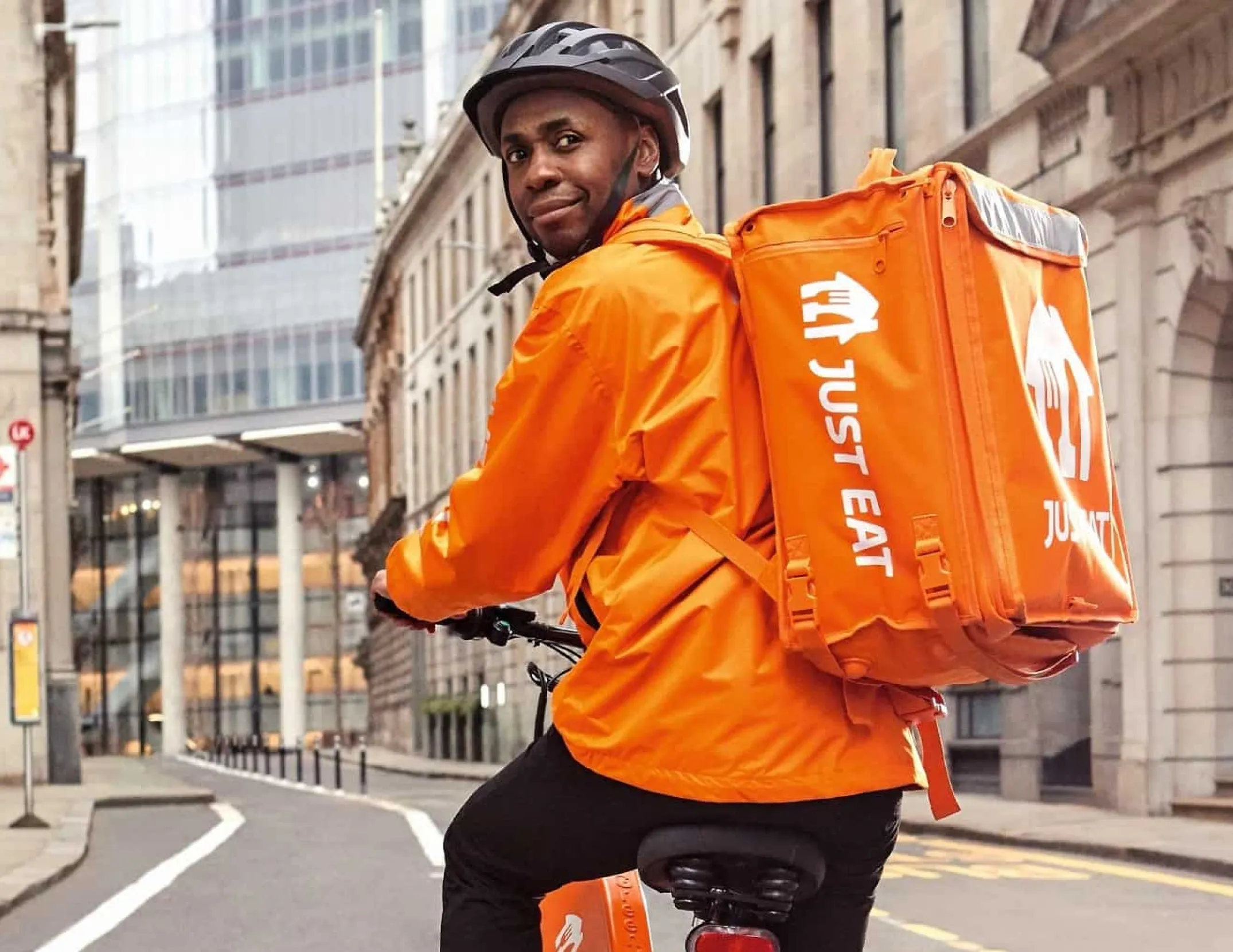 Man wearing black jacket riding a black Zoomo electric bike on a cobblestone street in front of a brick building.