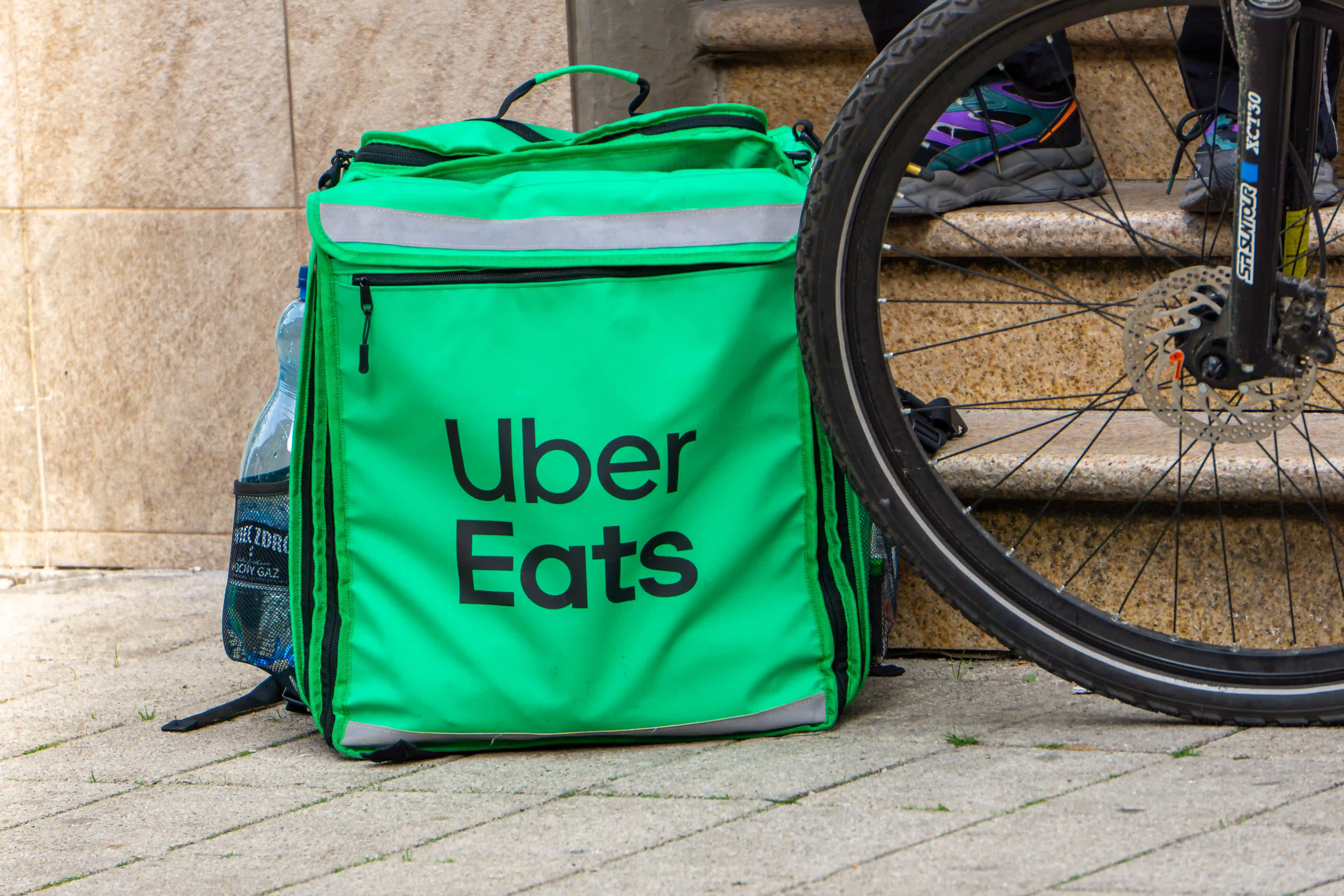 Man wearing black jacket riding a black Zoomo electric bike on a cobblestone street in front of a brick building.