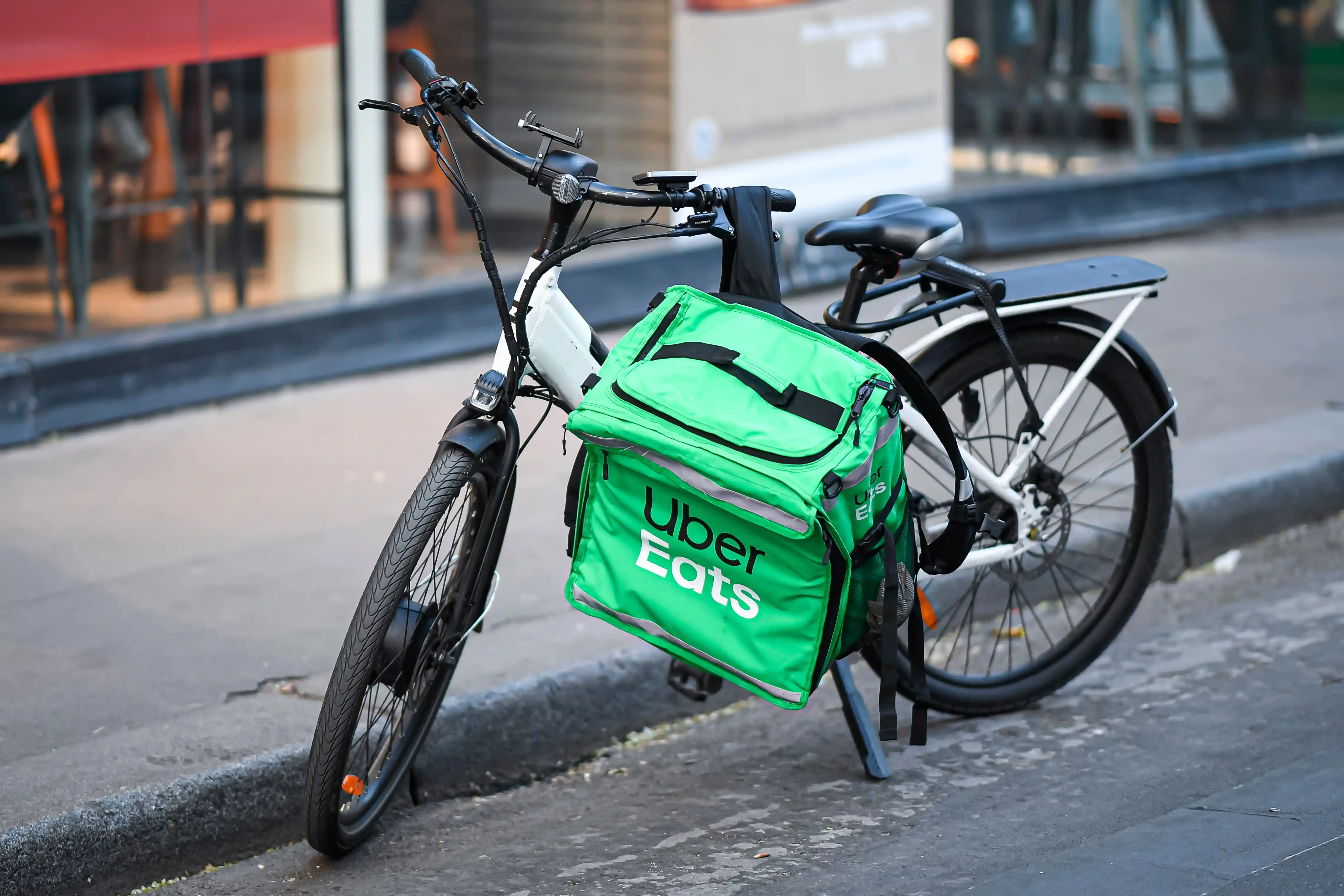 Woman in black jacket sitting on a white Zoomo electric bike with a large black backpack, in front of a brick and stone building.