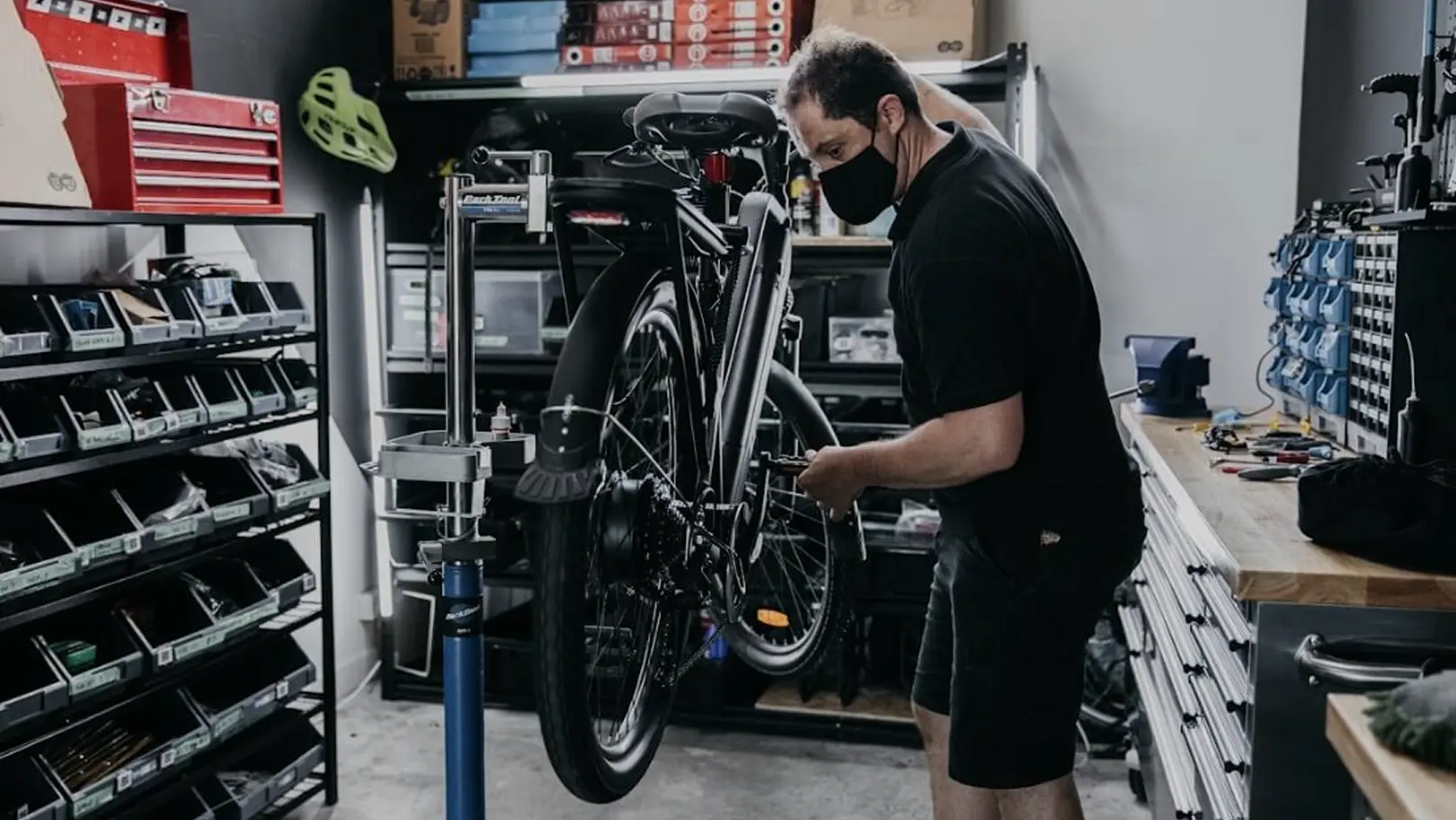 Man wearing a black face mask repairing a bicycle mounted on a repair stand in a workshop.