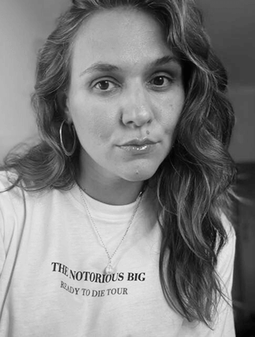 Black-and-white portrait of a woman with wavy hair wearing hoop earrings and a t-shirt reading 'The Notorious BIG Ready to Die Tour'.