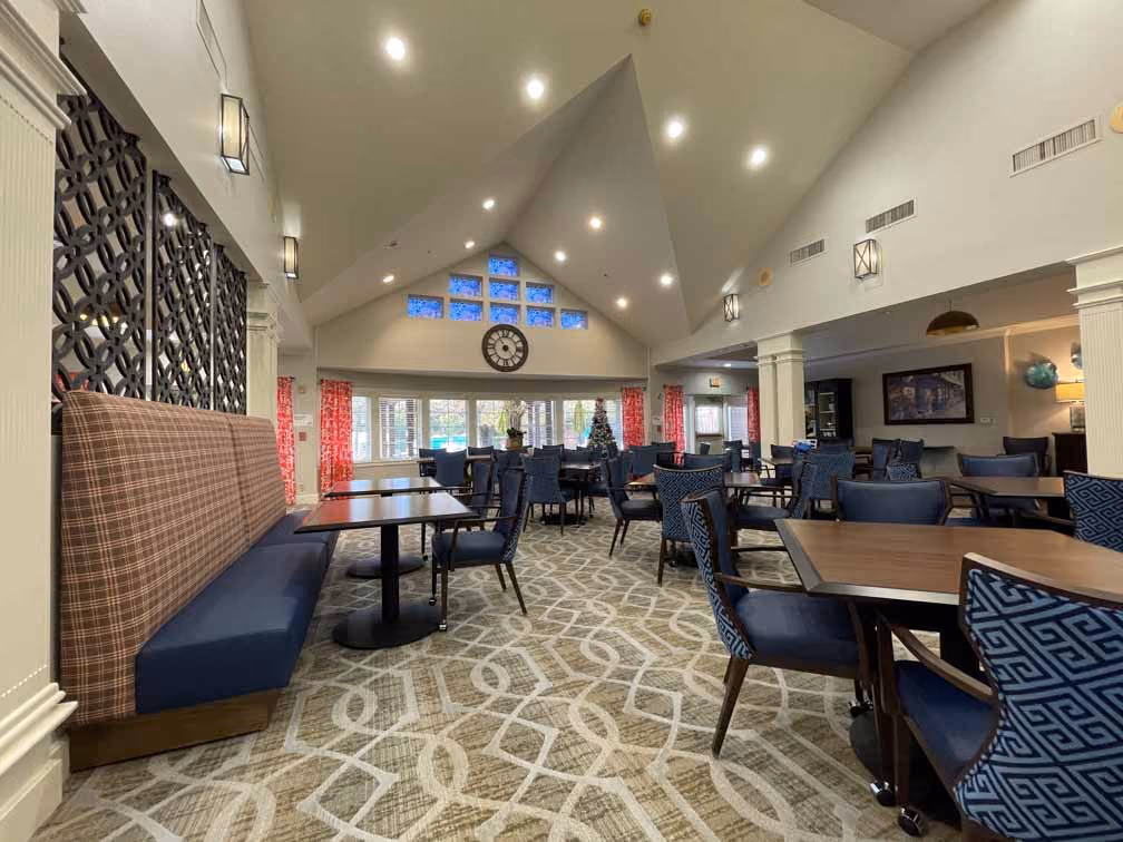 Spacious dining area with patterned carpet, plaid bench, blue upholstered chairs, wood tables, vaulted ceiling, and a large wall clock above windows.