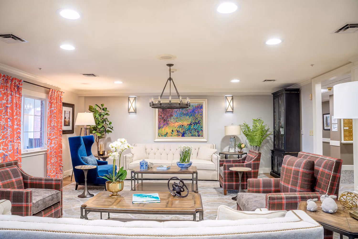 Well-lit modern living room with a beige tufted sofa, blue accent chair, plaid armchairs, colorful painting, and wooden coffee tables.