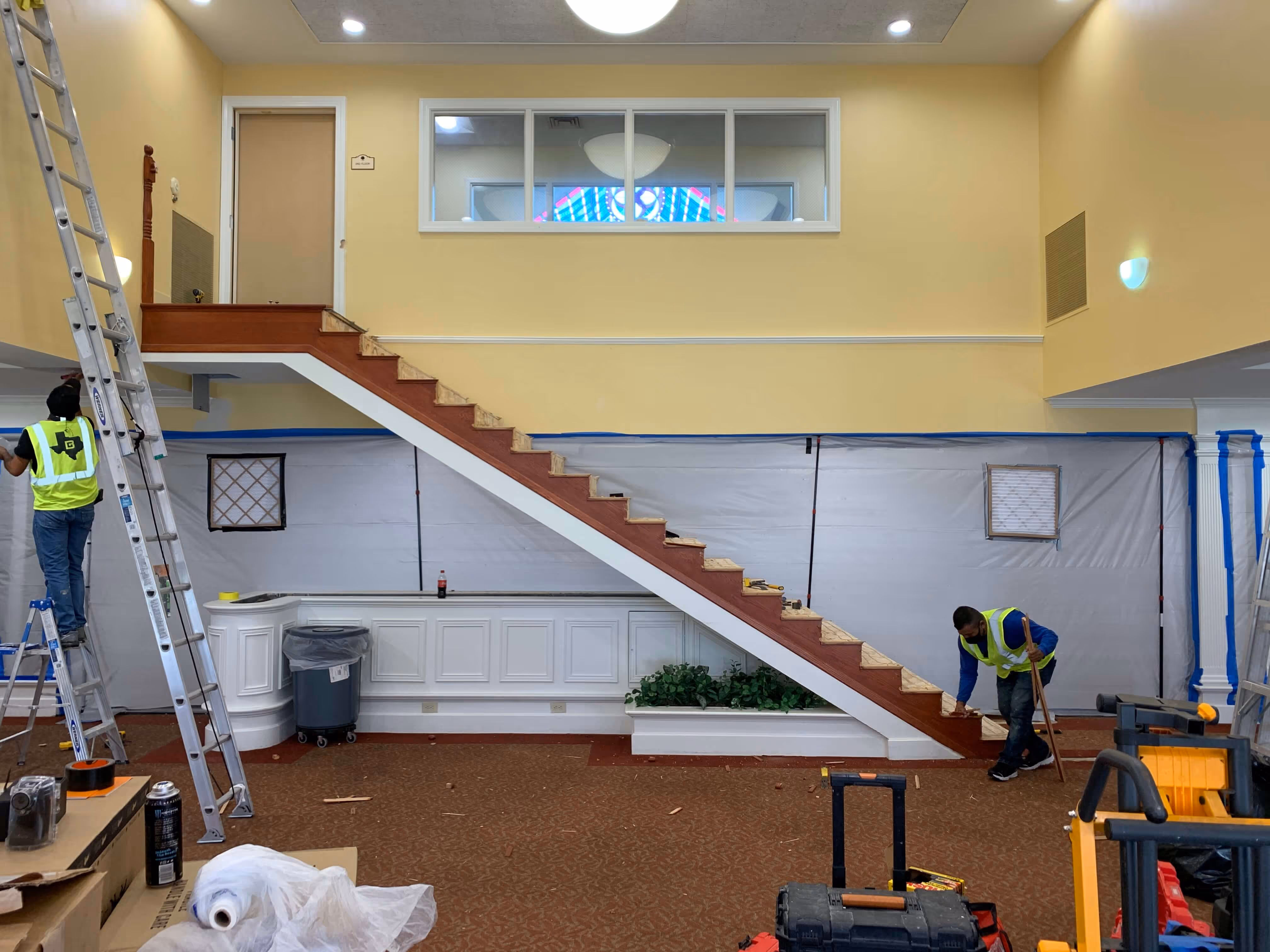 Two workers in neon vests renovating a room with a staircase, plastic-covered walls, and construction tools scattered around.