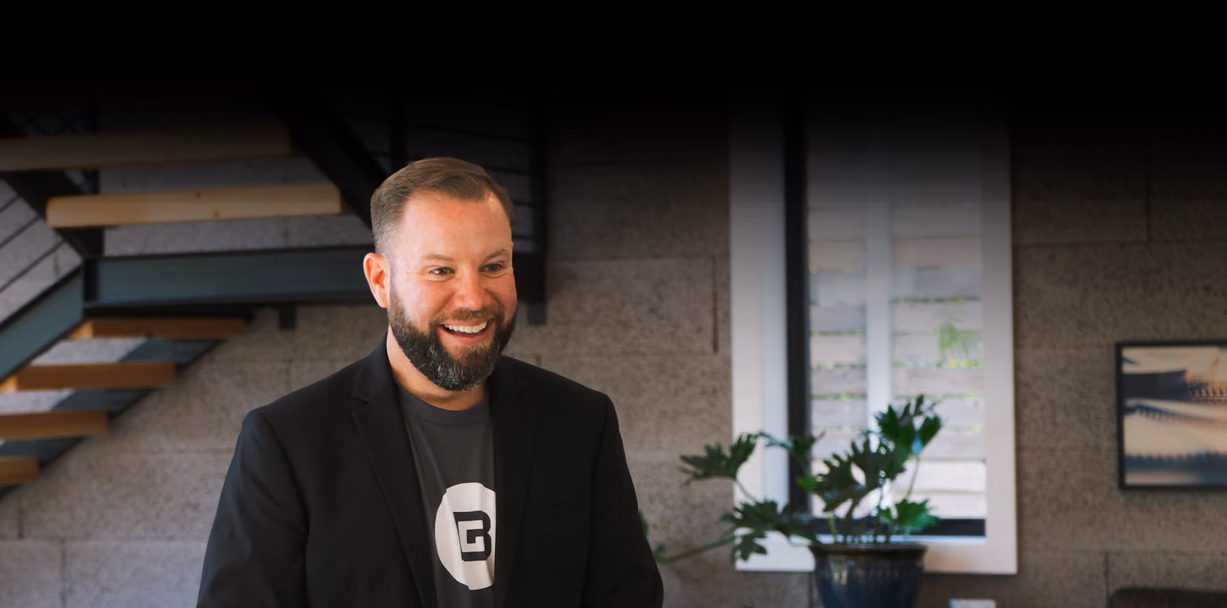 Smiling man with a beard wearing a black blazer and a grey t-shirt with a 'B' logo, standing indoors near a staircase and a window with a potted plant.