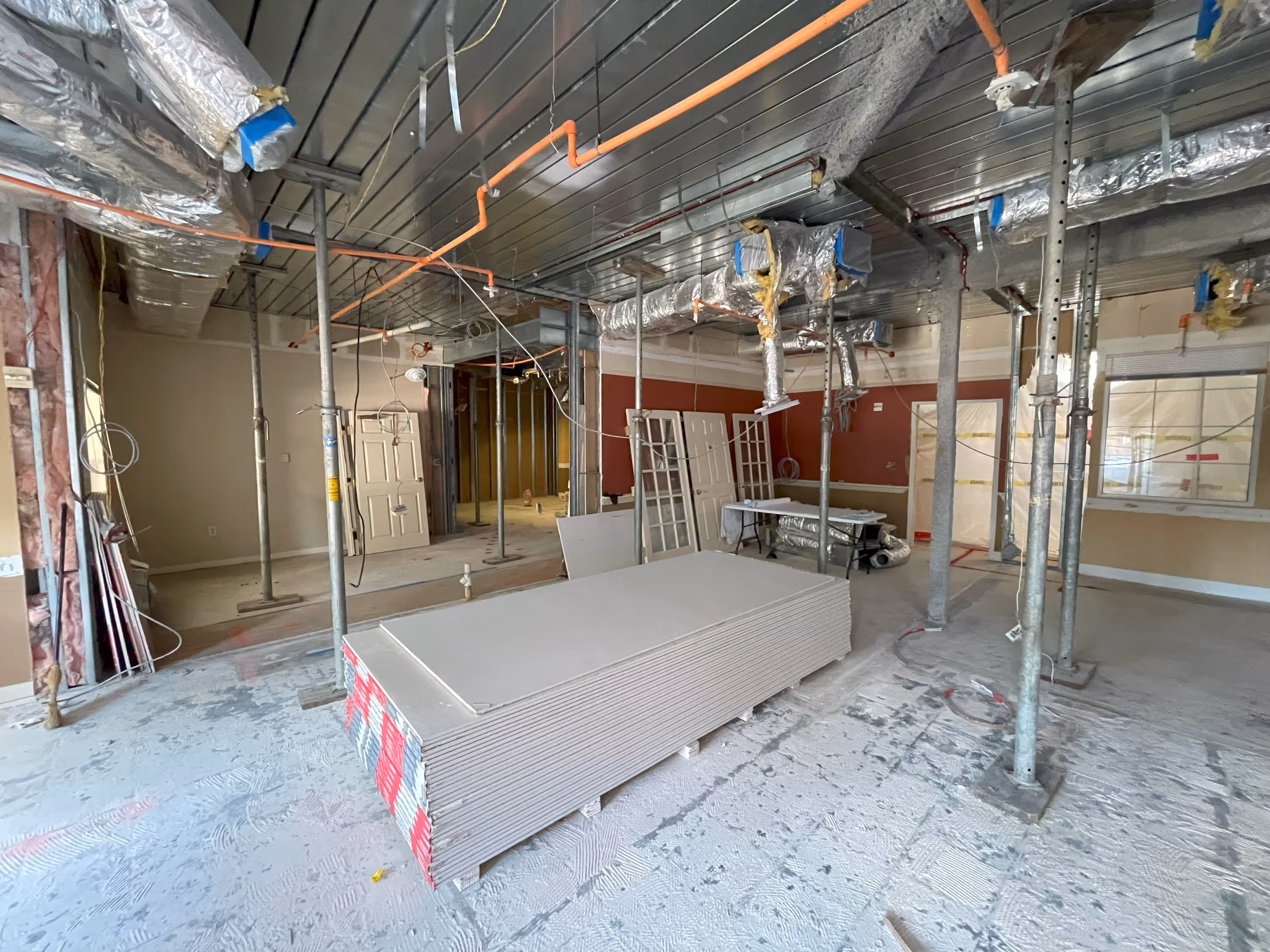 Interior of a construction site with drywall sheets stacked on the floor, exposed ceiling ducts wrapped in insulation, metal support poles, and various doors and windows leaning against walls.