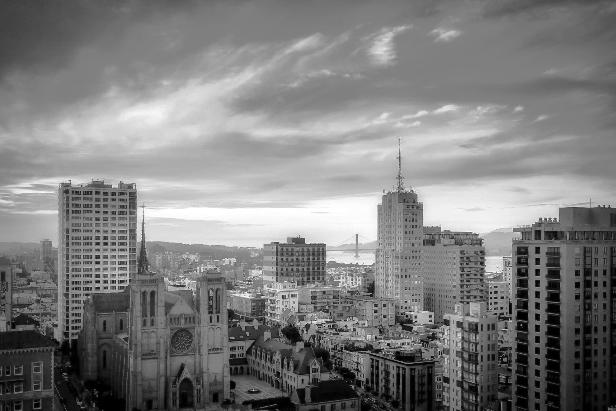 Black and white aerial view of a cityscape featuring a large Gothic-style cathedral, high-rise buildings, and a distant suspension bridge under a cloudy sky.