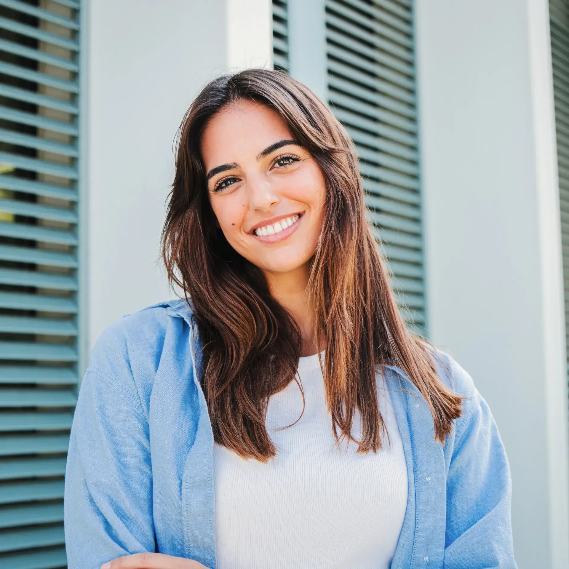 Smiling young woman with long brown hair wearing a white top and light blue shirt, standing outdoors near blue shutters.