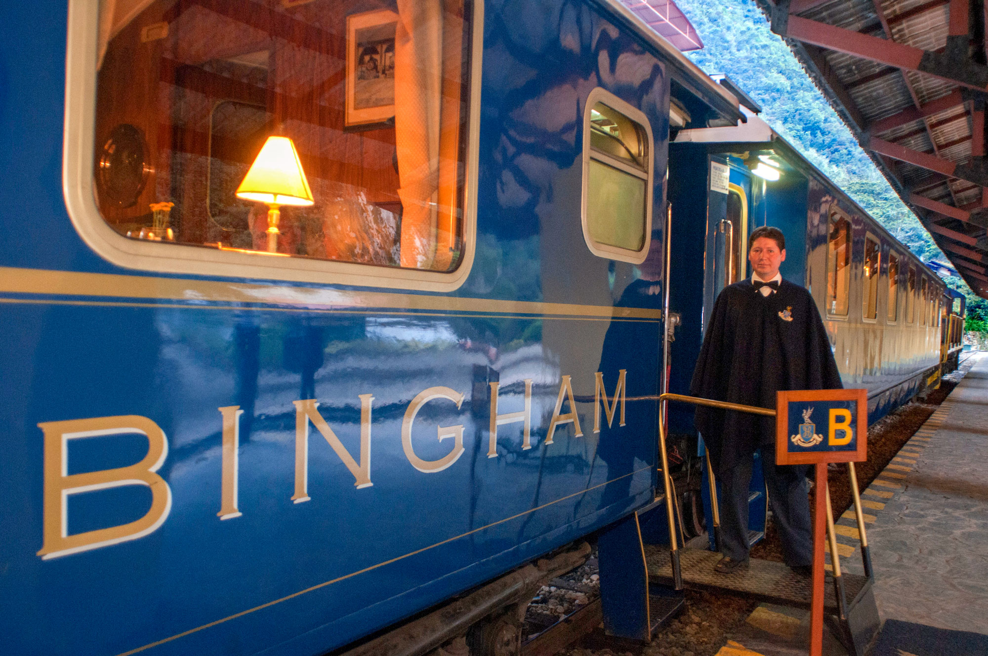 Close-up of the blue Belmond Hiram Bingham train car with a glowing interior lamp and a uniformed steward welcoming guests on the platform" caption="A warm Andean welcome: The glowing, elegant interiors of the Hiram Bingham await.