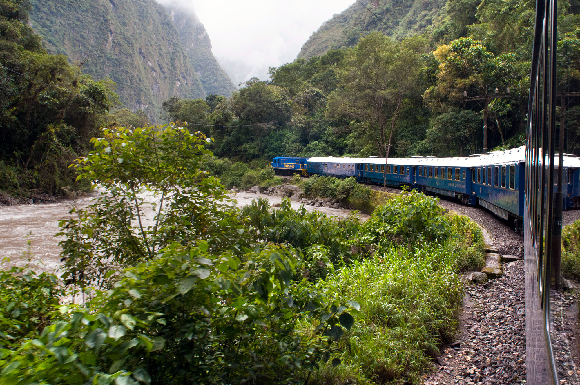 The luxury blue Hiram Bingham train curving along the tracks next to the rushing brown Urubamba River, surrounded by lush green mountain slopes and thick jungle vegetation in Peru.