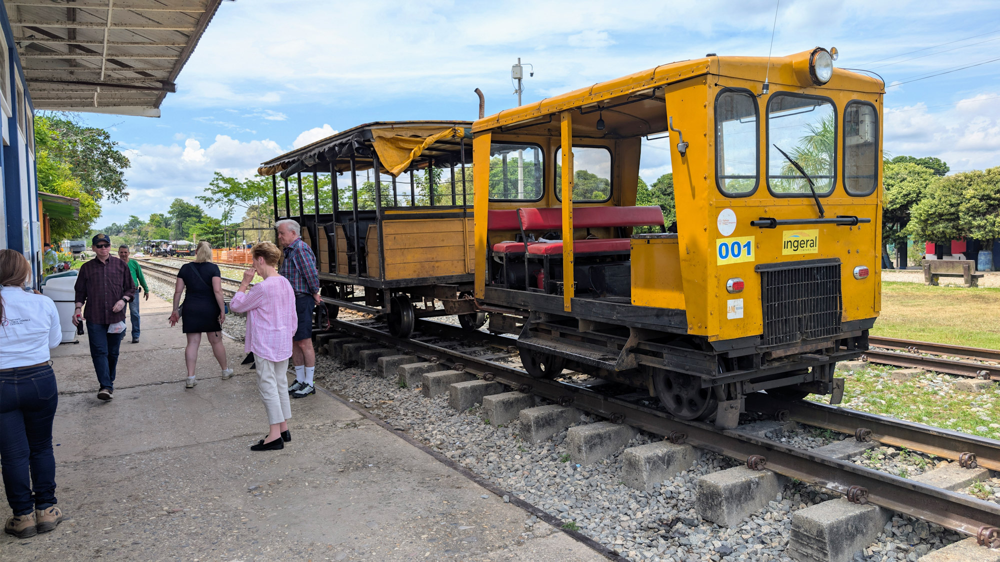 A group of tourists waiting on a concrete platform next to a yellow open-air carromotor rail vehicle at a Rio Magdalena railway station in Colombia.