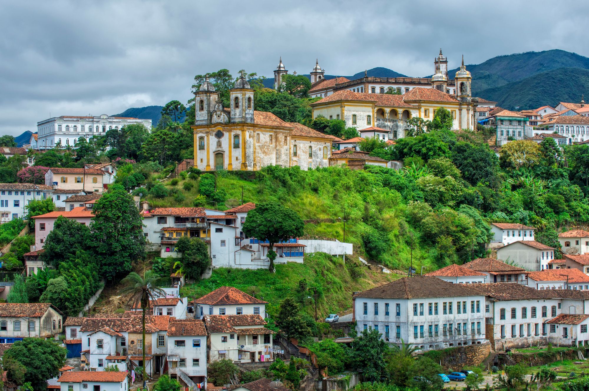 A scenic, high-angle view of the historic colonial town of Ouro Preto, Brazil, featuring whitewashed houses with orange terracotta tiled roofs nestled on a lush green hillside, with a yellow-and-white Baroque church standing prominently on a central hill under a cloudy sky and green mountains.