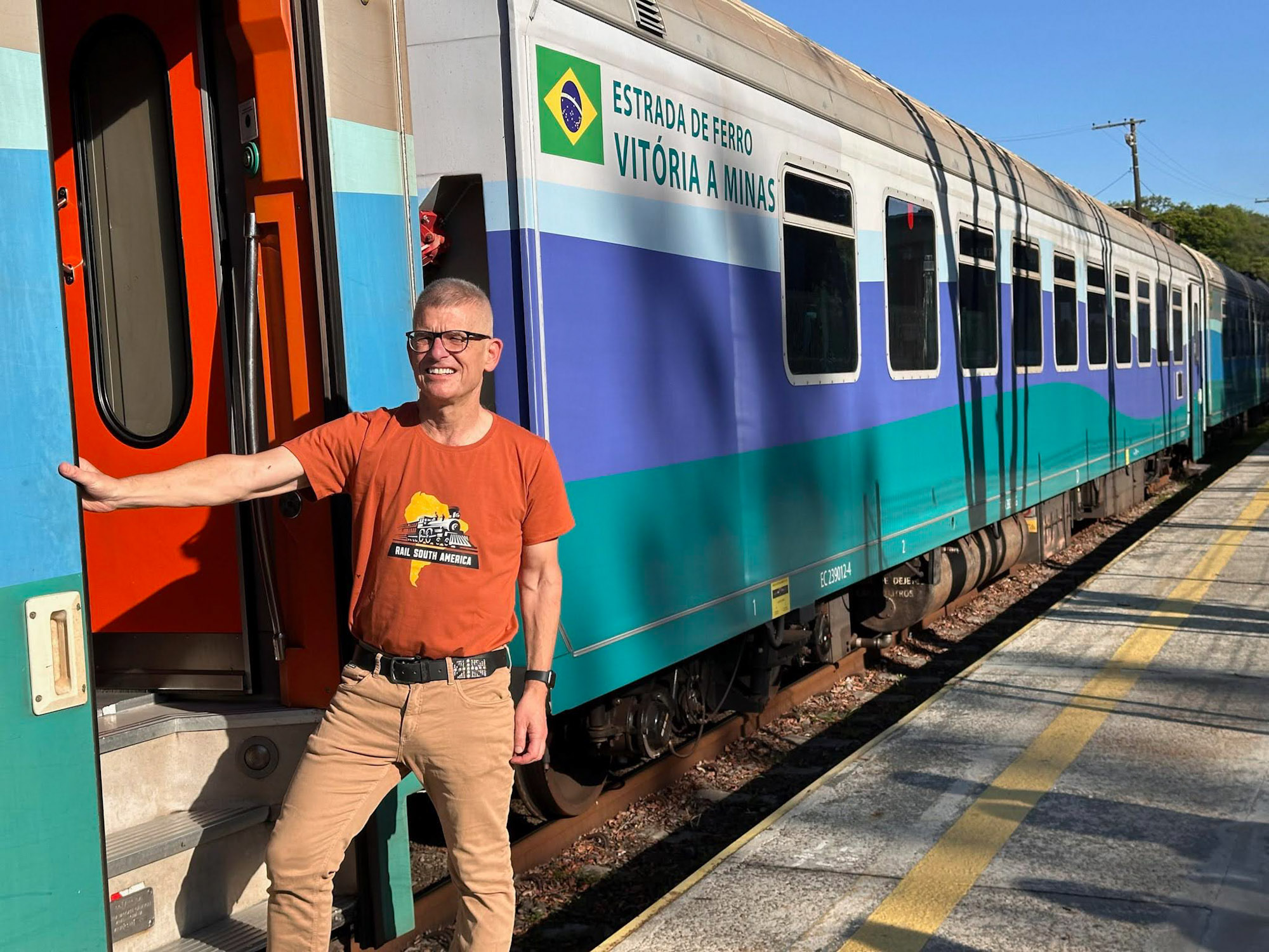 Peter Lauffer wearing an orange Rail South America t-shirt, smiling and boarding a blue, teal, and white passenger train carriage labeled "Estrada de Ferro Vitória a Minas" at the Vitória railway station on a sunny day.