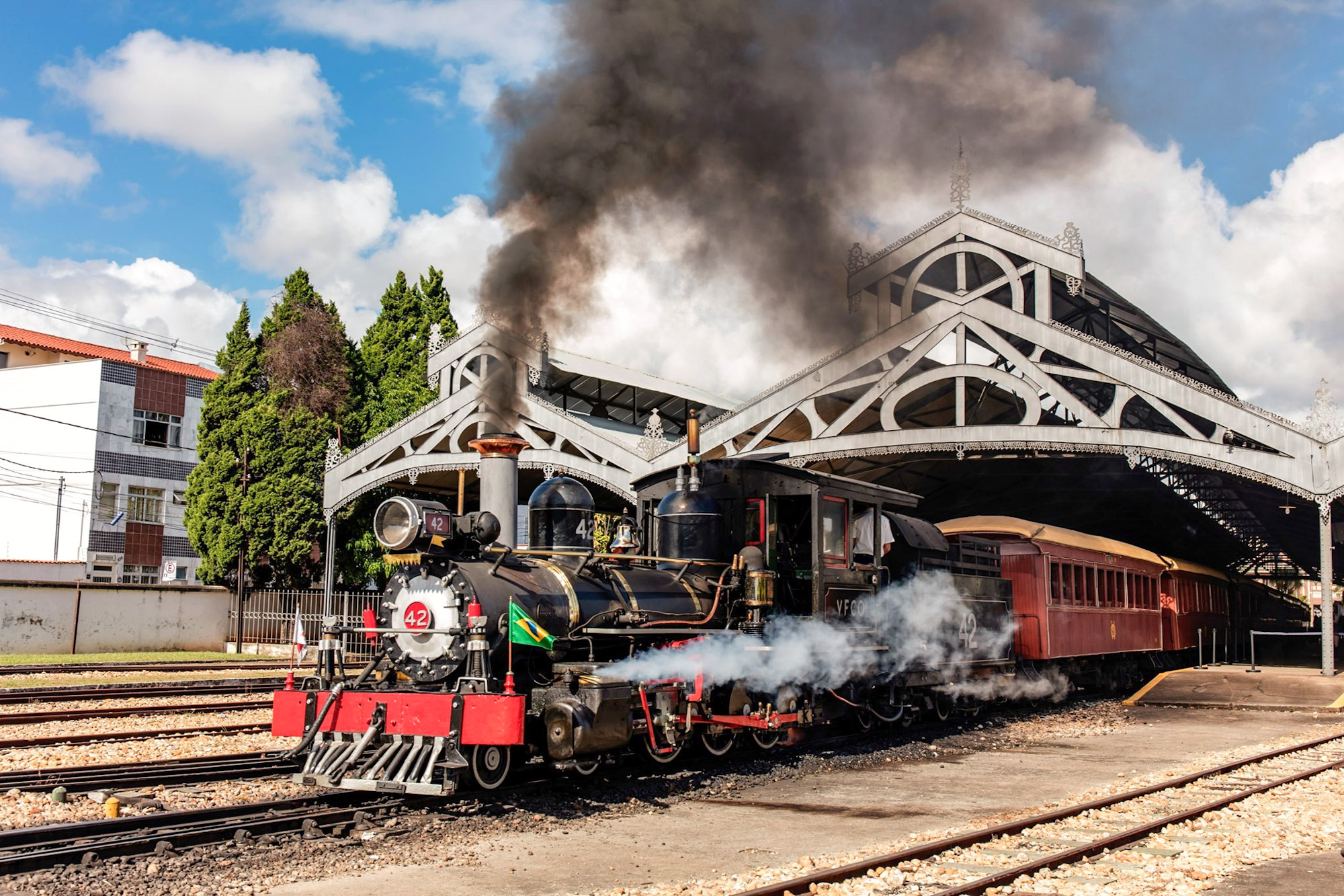 A historic black steam locomotive, number 42, billowing dark smoke and white steam as it sits under a large ornamental iron canopy at the São João del Rei railway station in Brazil with vintage passenger cars attached.