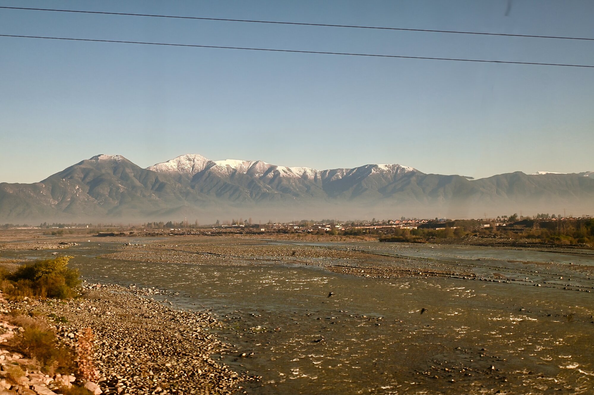Scenic view from a train window looking out over a wide river bed toward the snow-capped Andes Mountains in Chile's Central Valley.