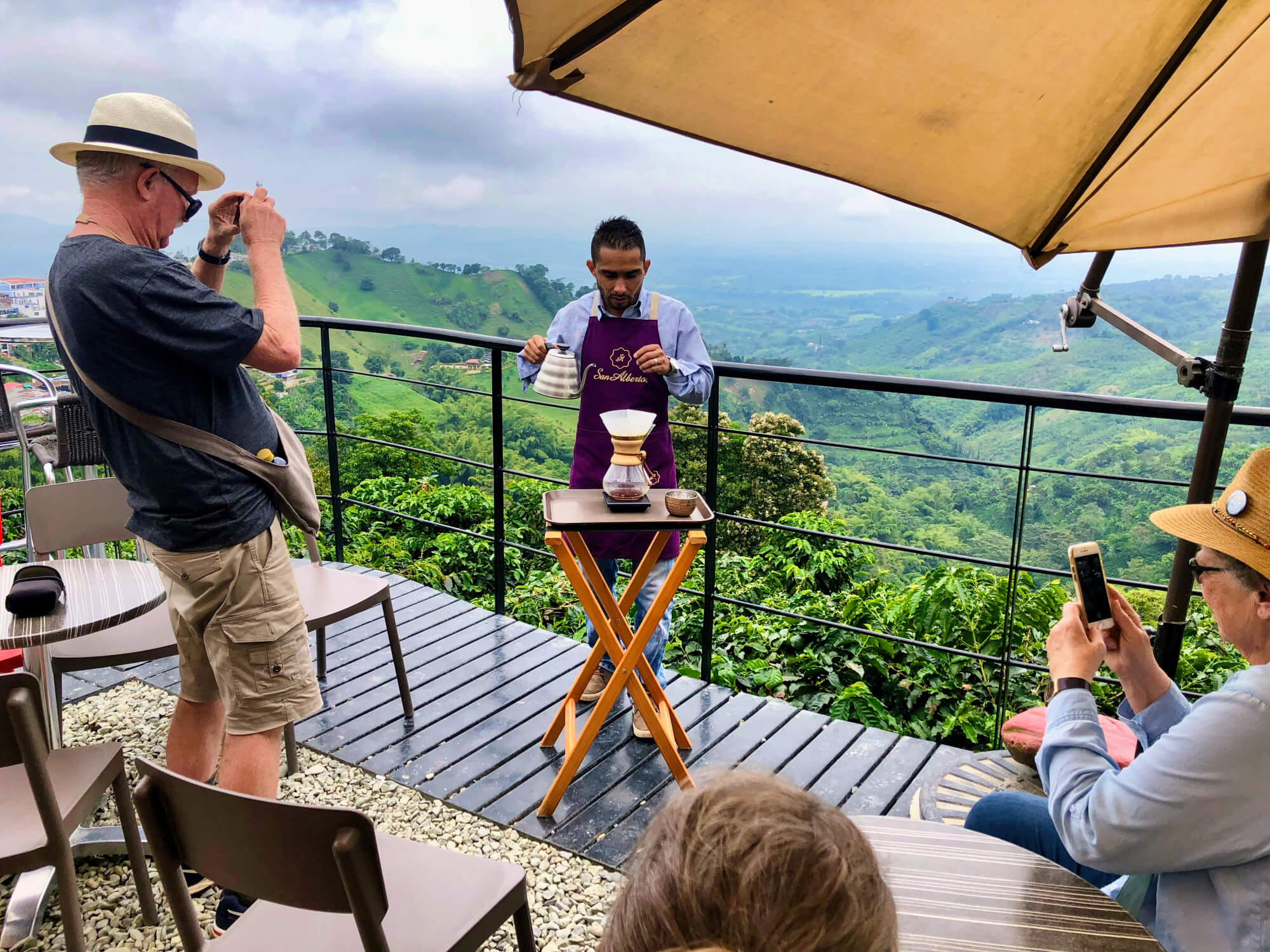 Coffee tasting demonstration on a terrace overlooking green mountain coffee country in Colombia, with travelers watching freshly brewed coffee being prepared.