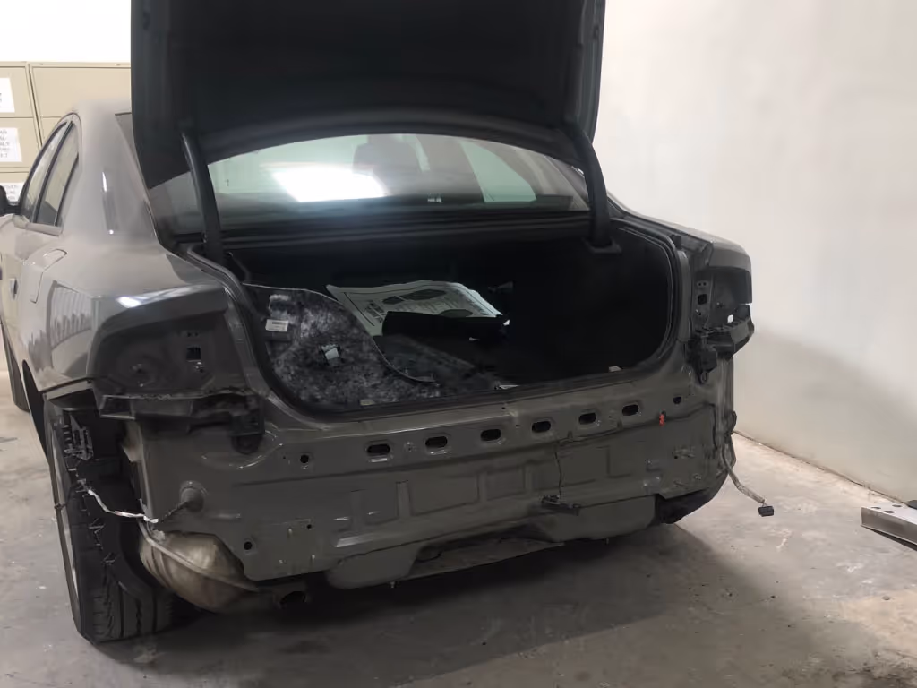 Car with open trunk missing its rear bumper and lights, parked indoors on a concrete floor.