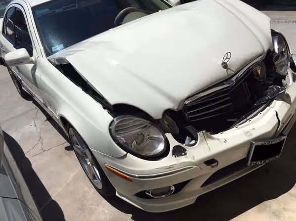 White Mercedes-Benz sedan with significant front-end damage, including a dented hood and broken grille and headlights.