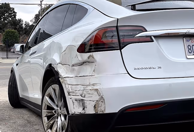 White Tesla Model X with significant damage and dents on the rear left side near the wheel.