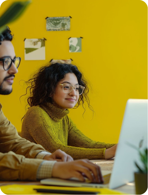 Two people with glasses working on laptops at a yellow-walled workspace.