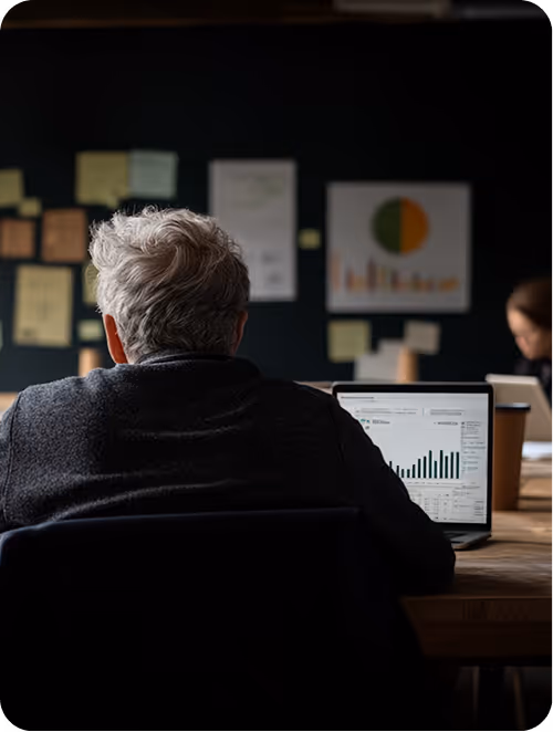 Two people with glasses working on laptops at a yellow-walled workspace.
