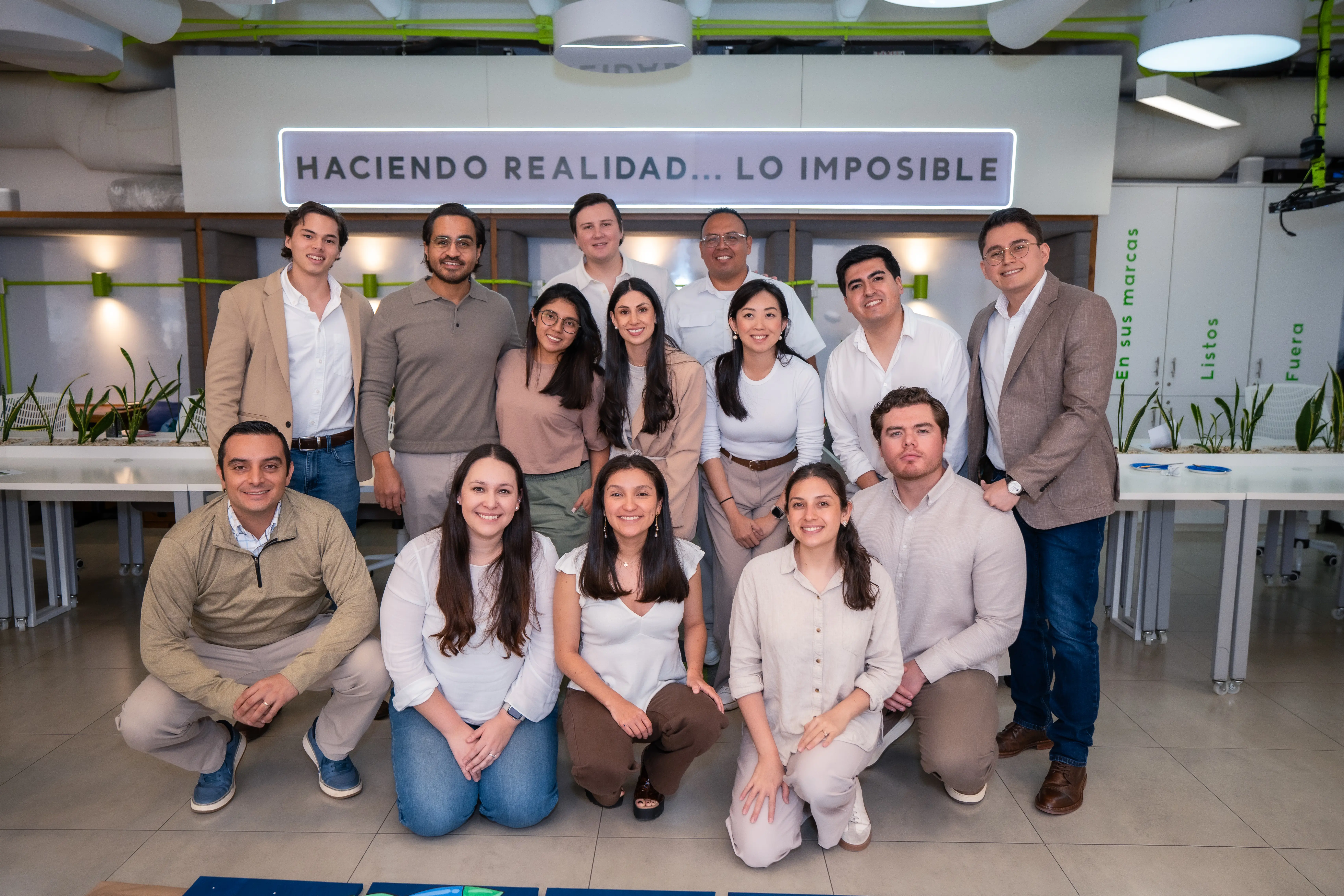 Group photo of fifteen smiling young adults in business casual attire posing indoors under a sign that reads 'Haciendo Realidad... Lo Imposible'.