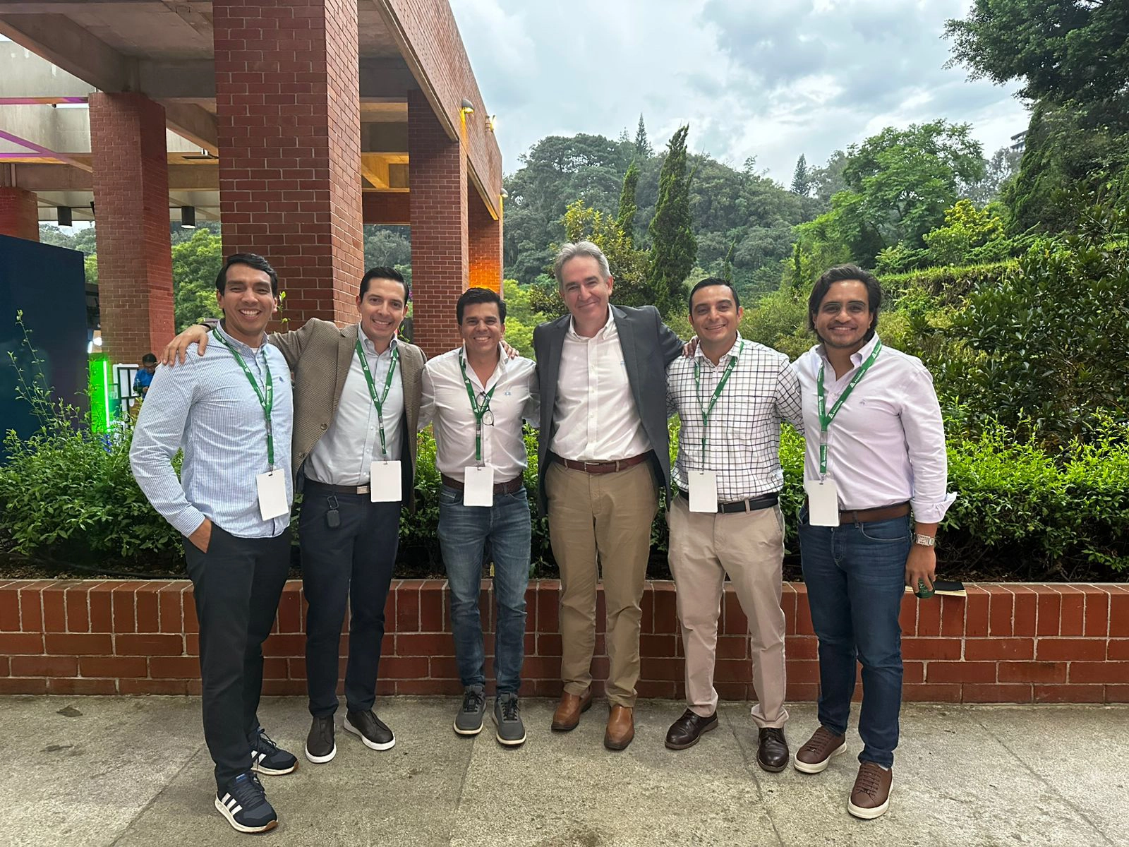 Group of six men standing arm-in-arm outdoors in front of brick planters and greenery, wearing business casual attire with green lanyards and badges.