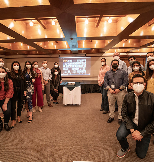 Group of people wearing masks standing in a conference room with a virtual meeting displayed on a projector screen behind them.