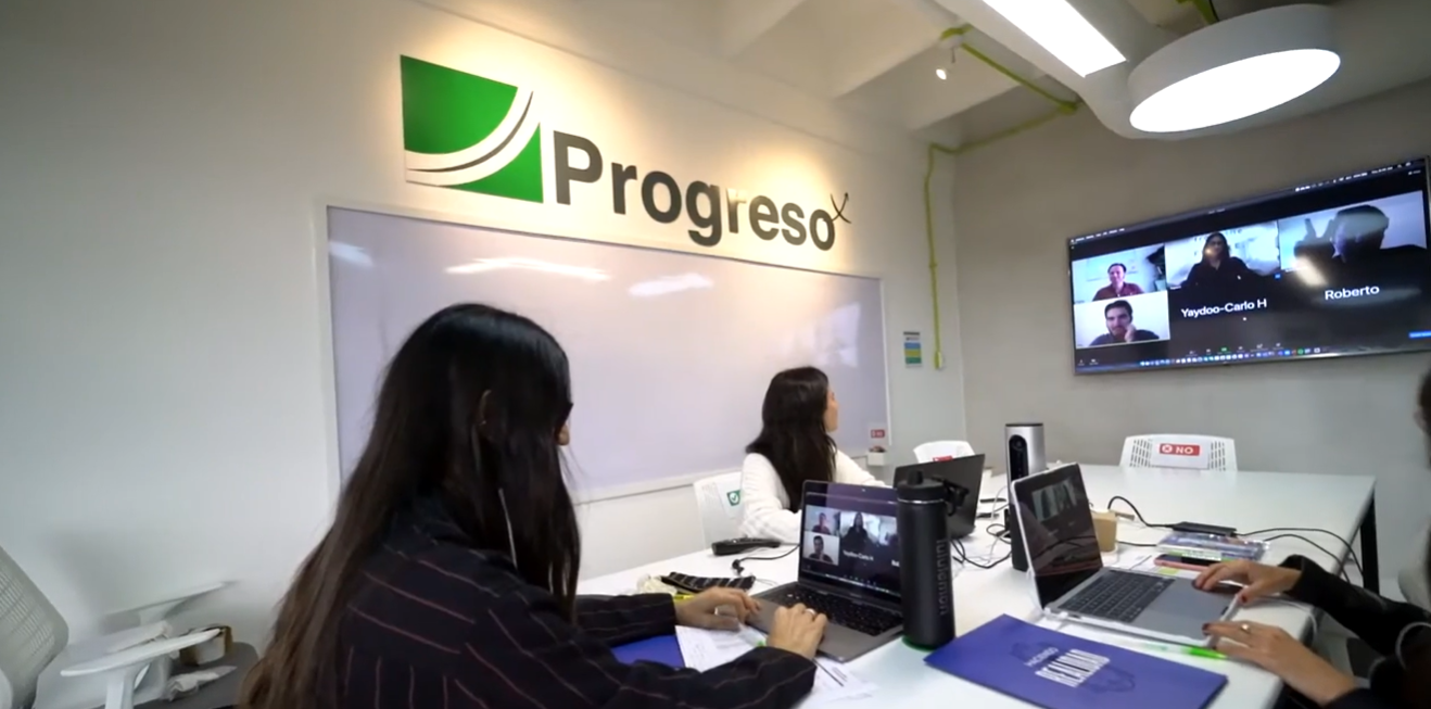Modern open office space with white desks and chairs, green plants dividing workstations, colorful carpet area, and ceiling lights with exposed pipes.