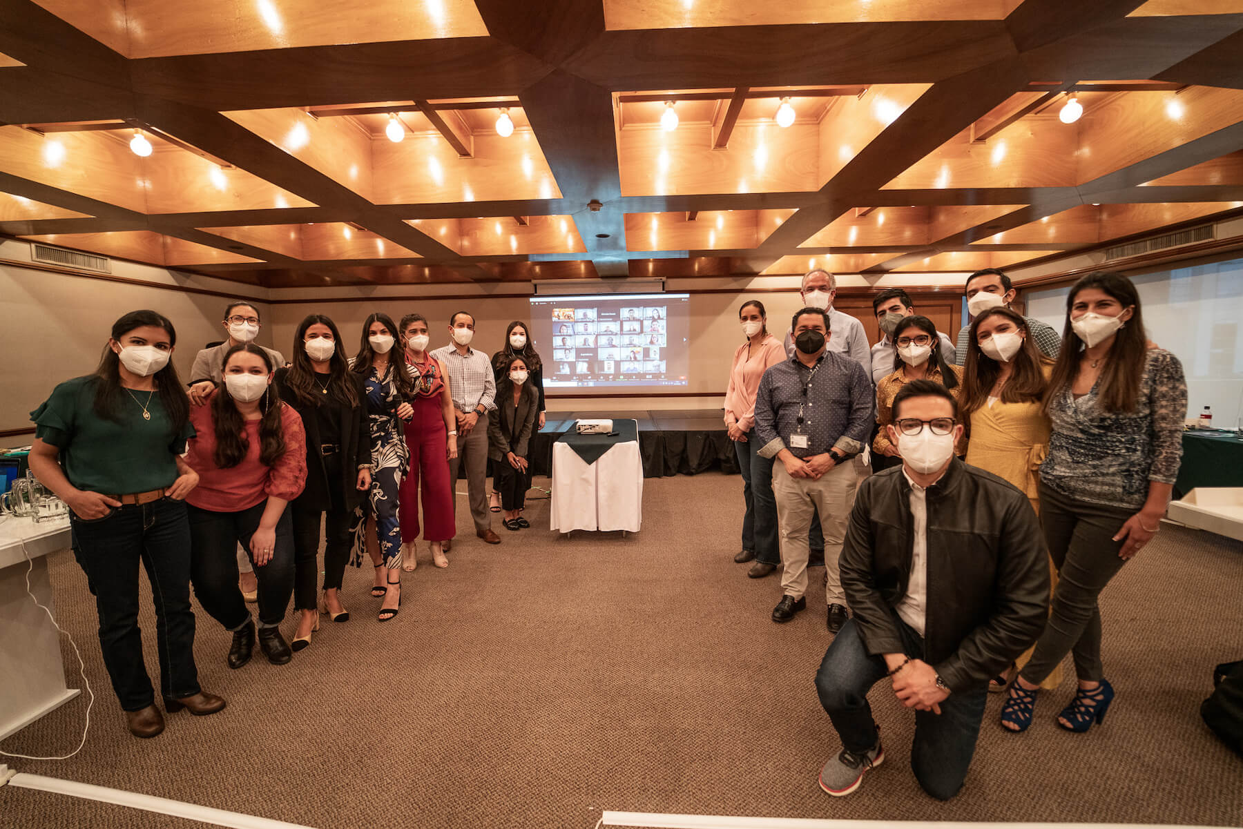 Modern open office space with white desks and chairs, green plants dividing workstations, colorful carpet area, and ceiling lights with exposed pipes.