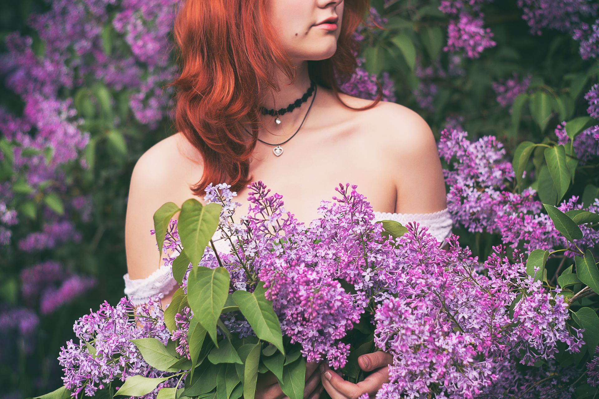 A woman with red hair holding vibrant lilacs, surrounded by lush greenery, exuding a feeling of spring.