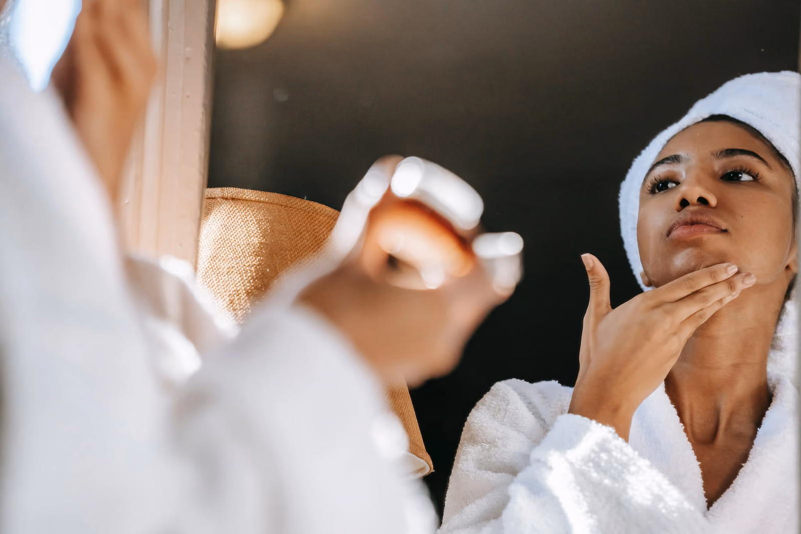Woman in a white bathrobe and towel applying skincare product to her chin while looking in the mirror.
